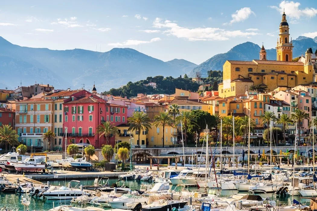Puerto costero pintoresco de la Riviera con casas tradicionales de colores, barcos anclados y una torre histórica sobre las montañas
